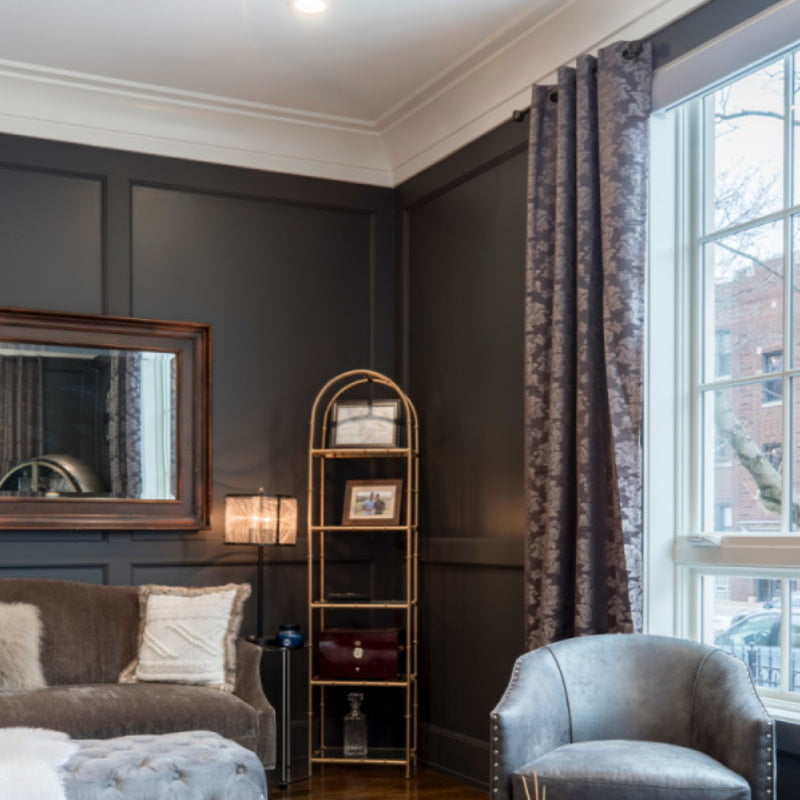 Elegant living room with dark wall paneling, gray furniture, crown molding, and large window draped with patterned curtains.