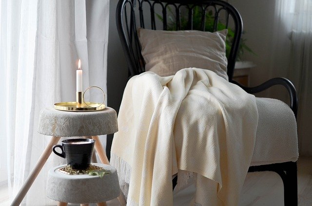 A cozy chair draped in a soft blanket beside a lit candle and coffee cup, creating a warm, tranquil reading nook.