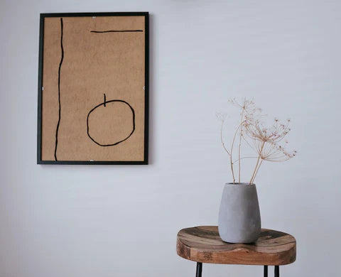 Abstract wall art in a black frame beside a wooden stool holding a concrete vase with dried flowers, embodying Japandi minimalism and natural textures.
